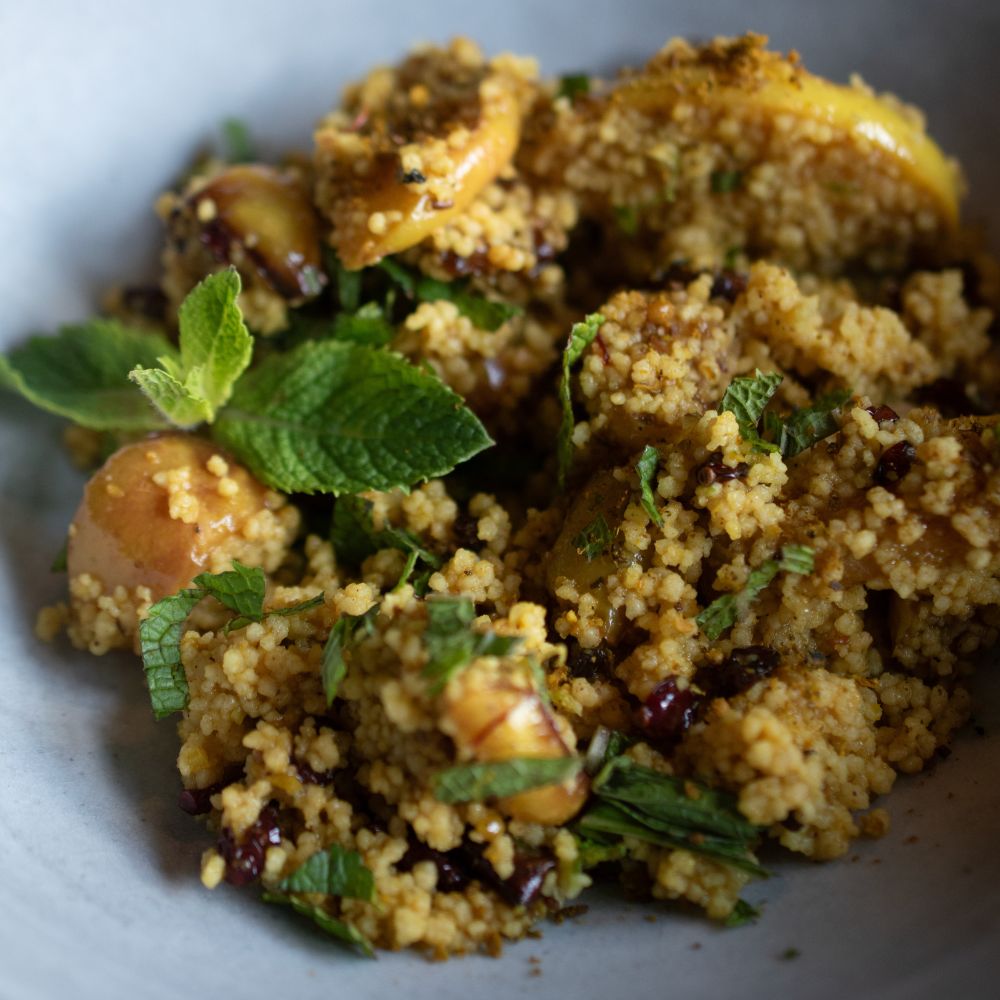 Fruchtiger Orangen-Couscous mit gebratenen Apfelspalten, Berberitzen und frischer Minze in einer hellen Keramikschüssel.