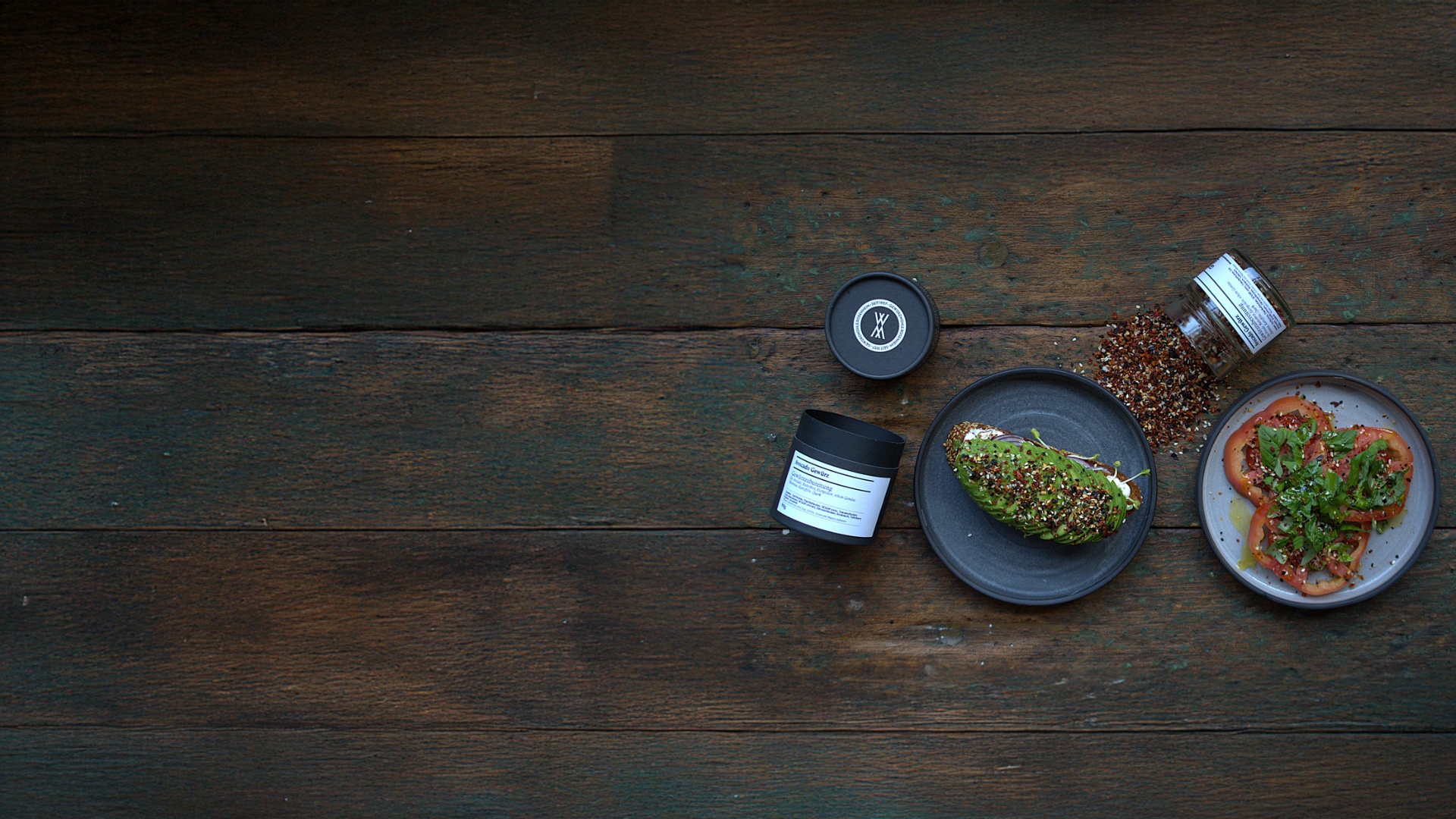 Dunkler Holztisch mit minimalistischer Food-Inszenierung: ein Teller mit Avocadobrot, daneben ein offenes Gewürzglas, ein zweiter Teller mit Tomaten und Basilikum sowie eine schwarze Gewürzdose – stilvolles Foodstyling mit Fokus auf Gewürze und Genuss