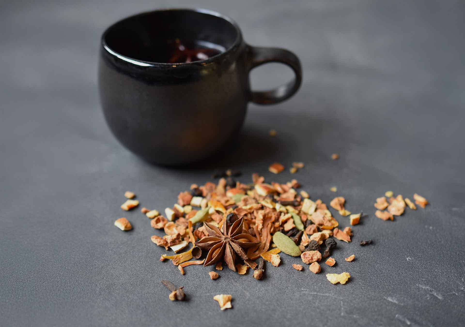 Eine schwarze Keramiktasse mit heißem Glühwein steht auf einem grauen Tisch. Daneben liegt lose Glühwein-Gewürzmischung mit Sternanis, Kardamom und Orangenschale – weihnachtliche Szene der Gewürzmühle Rosenheim.