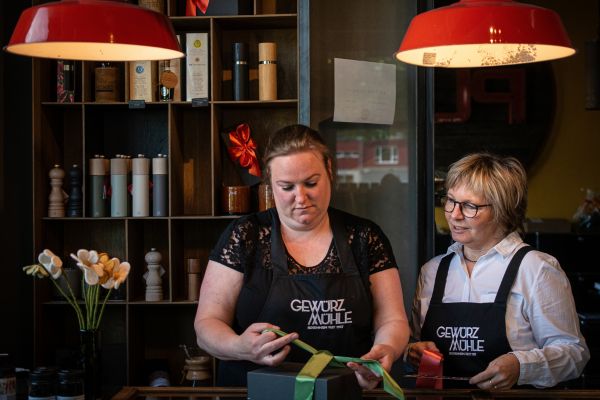Zwei Mitarbeiterinnen der Gewürzmühle Rosenheim stehen hinter dem Verkaufstresen und verpacken gemeinsam eine schwarze Geschenkbox mit grünem Geschenkband. Beide tragen schwarze Schürzen mit dem Logo der Gewürzmühle.