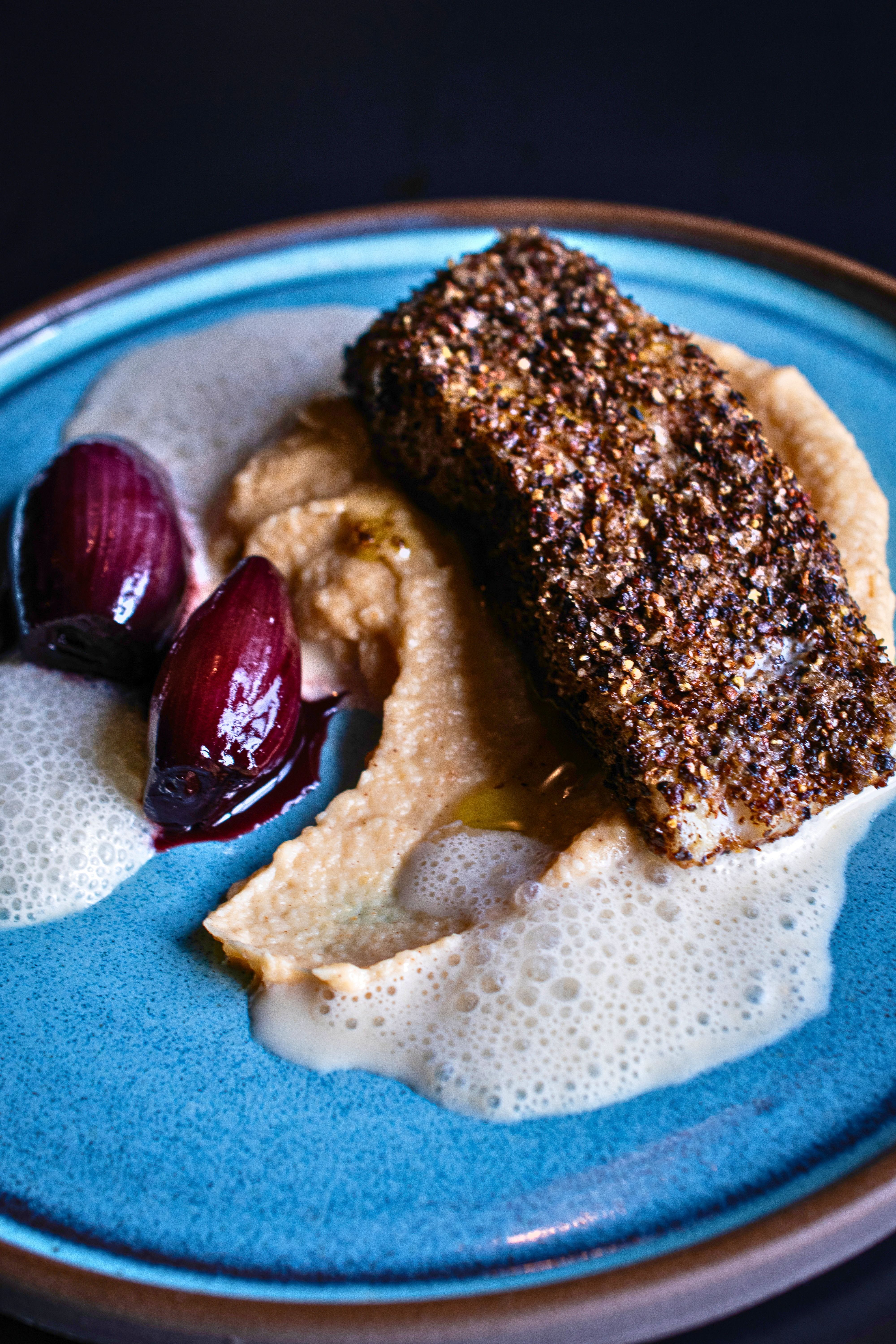 Nahaufnahme von Rotbarsch mit dunkler Pfefferkruste auf cremigem Püree, serviert mit glänzenden Portweinzwiebeln und einer aufgeschäumten Sauce auf einem blauen Teller.