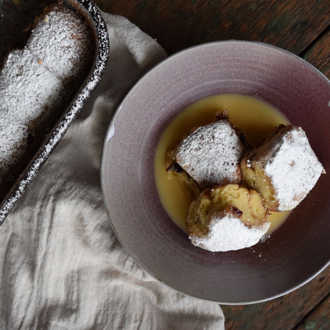Ein Dessertteller mit warmen Buchteln, bestäubt mit Puderzucker und serviert in Vanillesauce. Links daneben eine gebackene Form mit weiteren Buchteln. Rustikale Präsentation auf Holzoberfläche im Stil der Gewürzmühle Rosenheim.