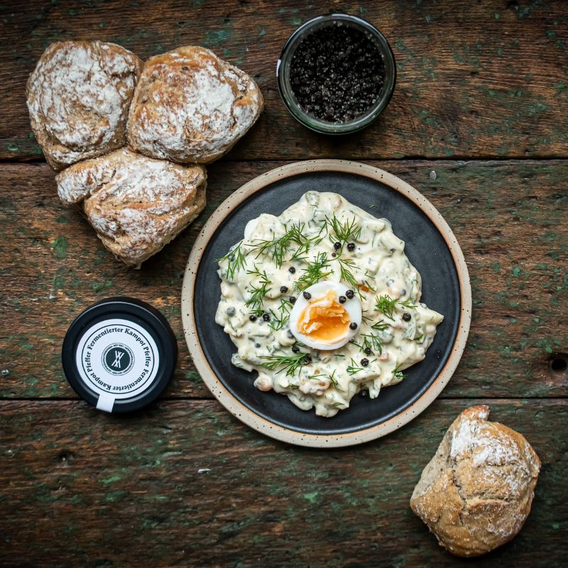 Eiersalat mit Dill und halbiertem Ei auf einem Teller, umgeben von Brötchen und einem Glas fermentierter Kampot Pfeffer der Gewürzmühle Rosenheim. Rustikaler Holzhintergrund betont die natürliche Rezeptidee.