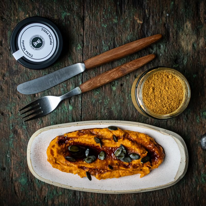 Kürbisaufstrich mit Tikka-Masala-Gewürzmischung und Kürbiskernen auf Keramikteller. Daneben Glas und Dose der Gewürzmühle Rosenheim mit der Mischung „Tikka Masala“, sowie Besteck aus Holzgriff.
