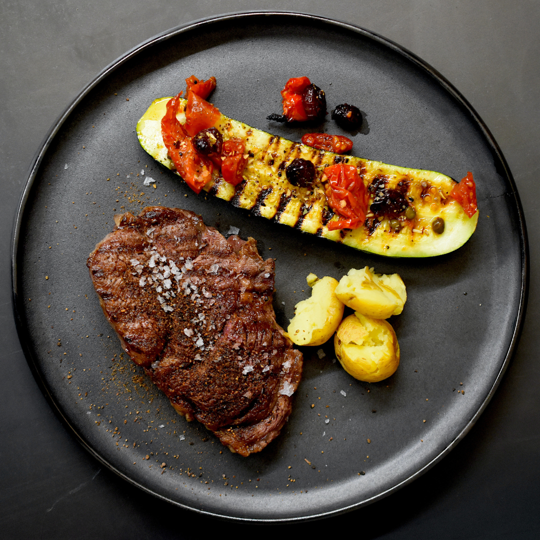 Steak, Kartoffeln und Zucchinischeibe auf dunklem Teller serviert, auf dunklem Untergrund