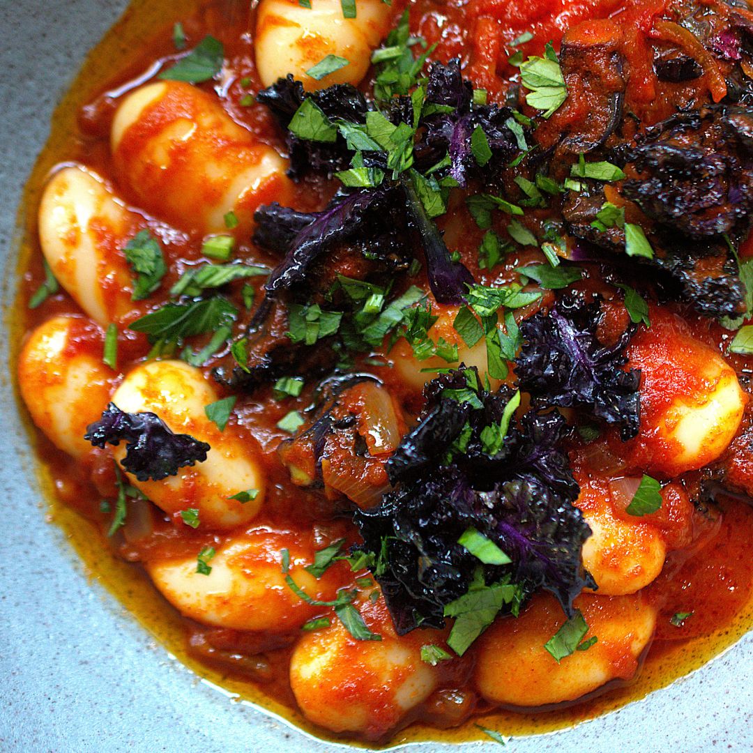 Nahaufnahme Butterbohnen in Tomatensauce und Grünkohlsprossen sowie Kräutern in einem tiefen Teller.