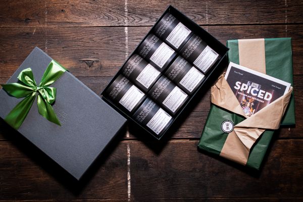 Verschiedene Geschenkideen der Gewürzmühle Rosenheim auf einem rustikalen Holztisch: ein geöffnetes Set mit acht schwarzen Gewürzdosen sowie ein in grünes Papier verpacktes Geschenk mit dem Kochbuch SPICED.