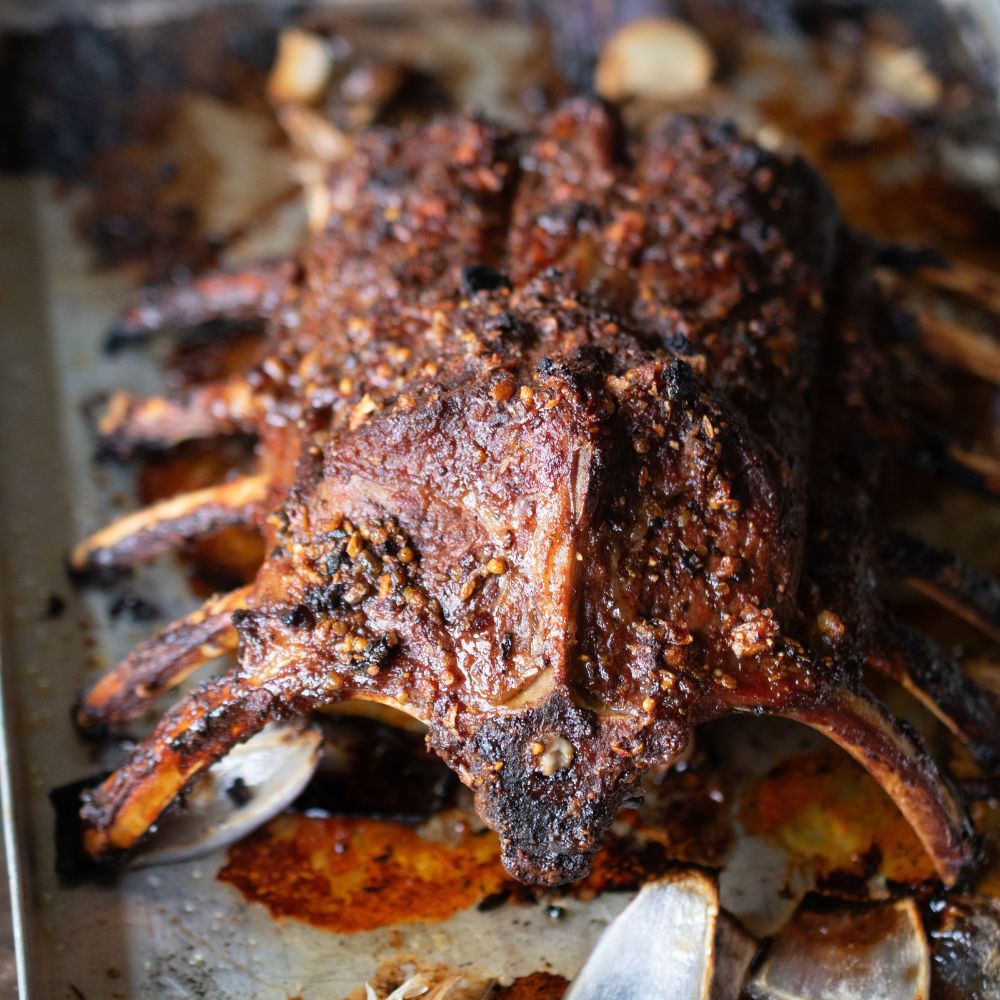Knusprig gebratenes Lammkarree mit kräftiger Gewürzkruste, gerösteten Zwiebeln und dunklem Bratensaft auf einem Backblech.