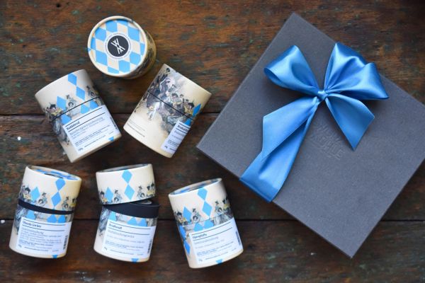 Fünf Gewürzdosen der Gewürzmühle Rosenheim mit blau-weißem Rautenmuster und historischen Illustrationen liegen neben einer schwarzen Geschenkbox mit großer blauer Schleife auf einem rustikalen Holztisch.