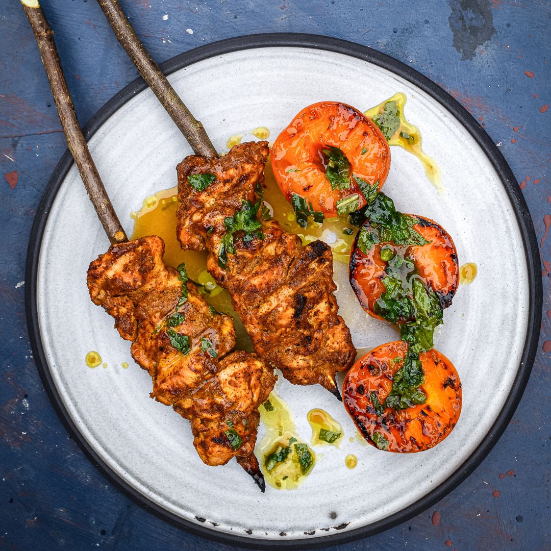 Zwei kräftig gewürzte Fleischspieße mit gegrillten Aprikosenhälften und Kräuteröl auf hellem Teller. Zubereitet mit Gewürzen der Gewürzmühle Rosenheim.