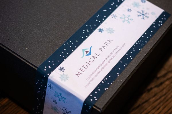 Nahaufnahme einer schwarzen Geschenkbox mit einer personalisierten Banderole für Medical Park. Die weiß-dunkelblaue Banderole ist mit Schneeflocken und winterlichen Motiven gestaltet.