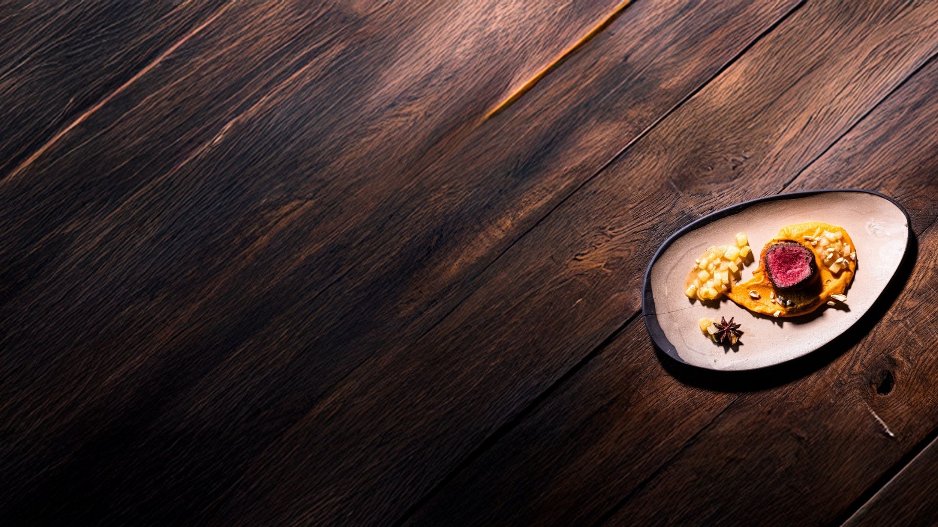 Holzuntergrund mit einem Teller, auf dem ein Stück Rehrücken auf Süsskartoffelcreme gewürzt mit Five Spices liegt.