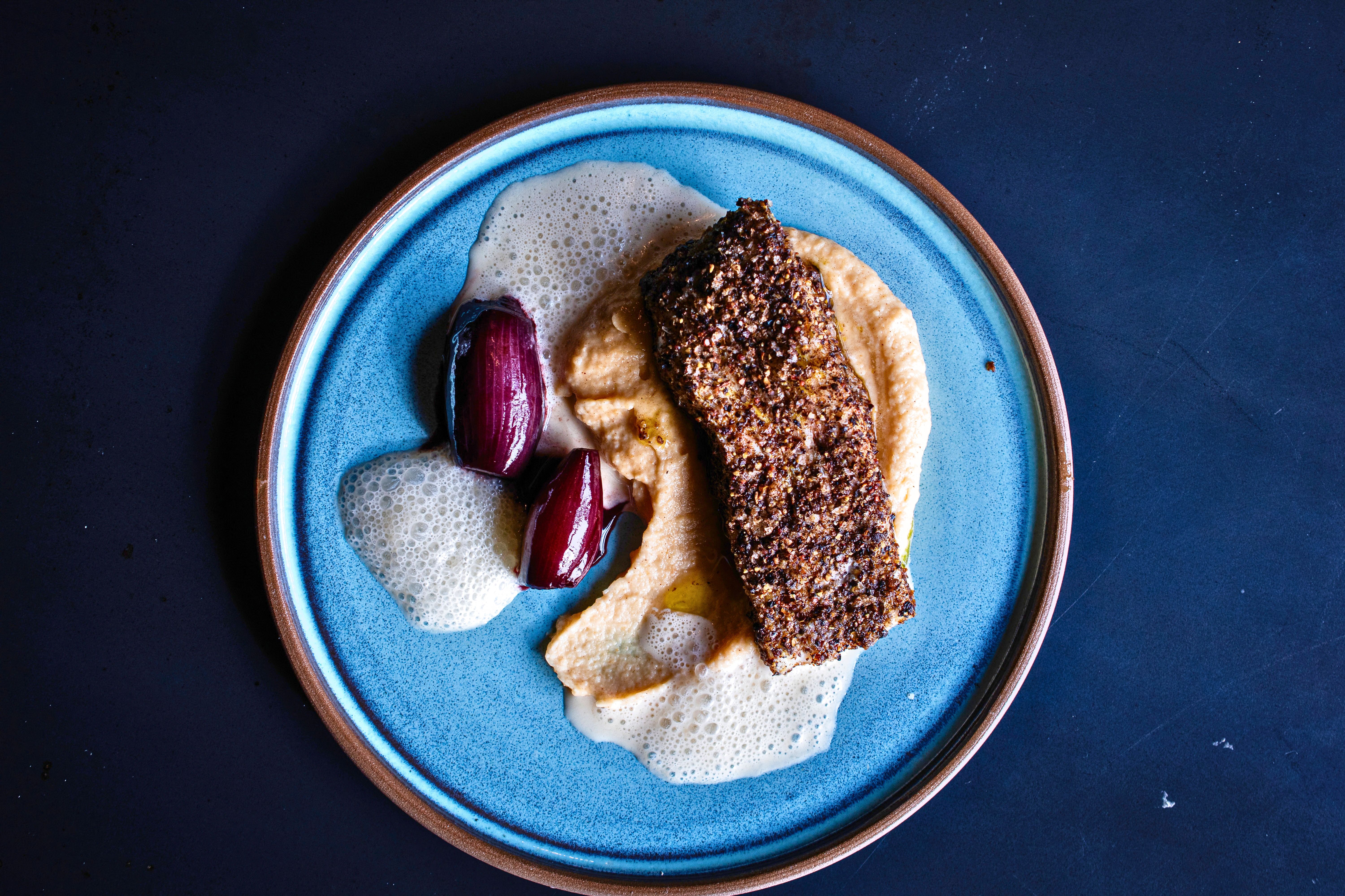Rotbarsch mit kräftiger Pfefferkruste, cremigem Püree und Portweinzwiebeln auf einem blauen Keramikteller. Eine helle, aufgeschäumte Sauce rundet das angerichtete Fischgericht ab.
