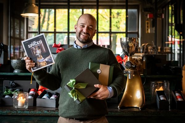 Ein lächelnder Mann im grünen Pullover hält das Kochbuch SPICED und eine schwarze Geschenkbox mit grüner Schleife der Gewürzmühle Rosenheim in den Händen. Im Hintergrund stehen weitere Geschenksets und eine goldene Gewürzmühle.