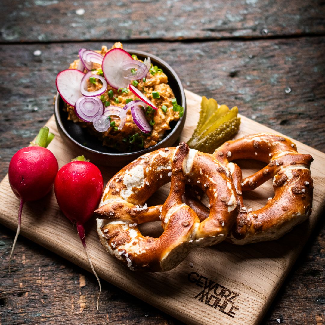 Eine bayerische Brotzeit auf einem Holzbrett der Gewürzmühle Rosenheim: Obazda in einer kleinen Schale, dekoriert mit Radieschen und Zwiebeln, dazu frische Brezeln und eingelegte Gurken. Alles arrangiert auf dunklem Holz.