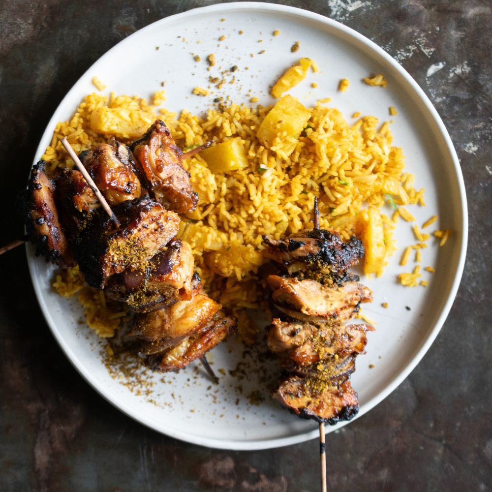 Gegrillte Hähnchenspieße mit gelbem Curryreis und Ananasstücken auf einem großen hellen Teller.