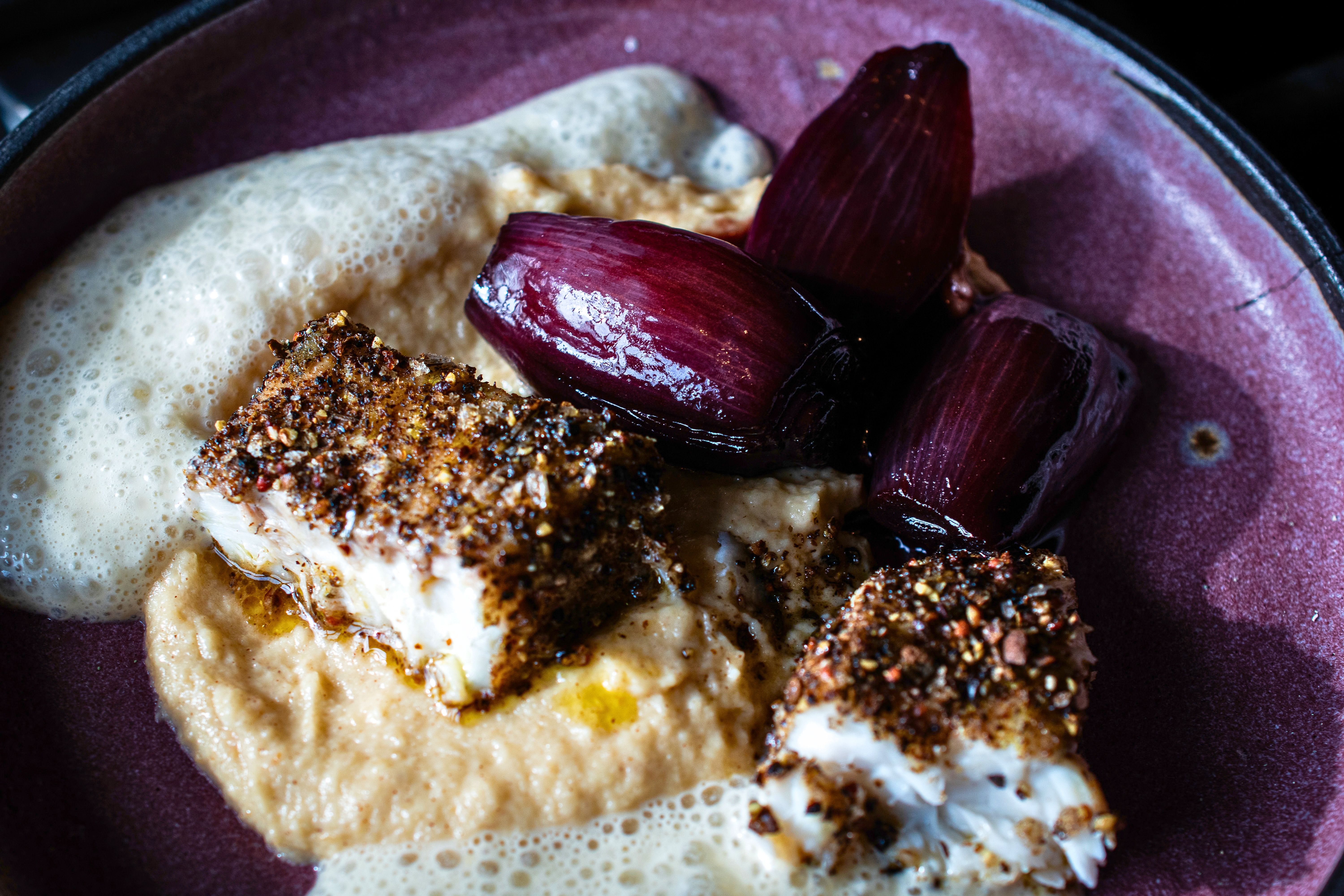 Detailaufnahme von aufgeschnittenem Rotbarsch mit würziger Pfefferkruste, cremigem Püree und dunkelroten Portweinzwiebeln. Dazu ist eine helle, fein aufgeschäumte Sauce angerichtet.