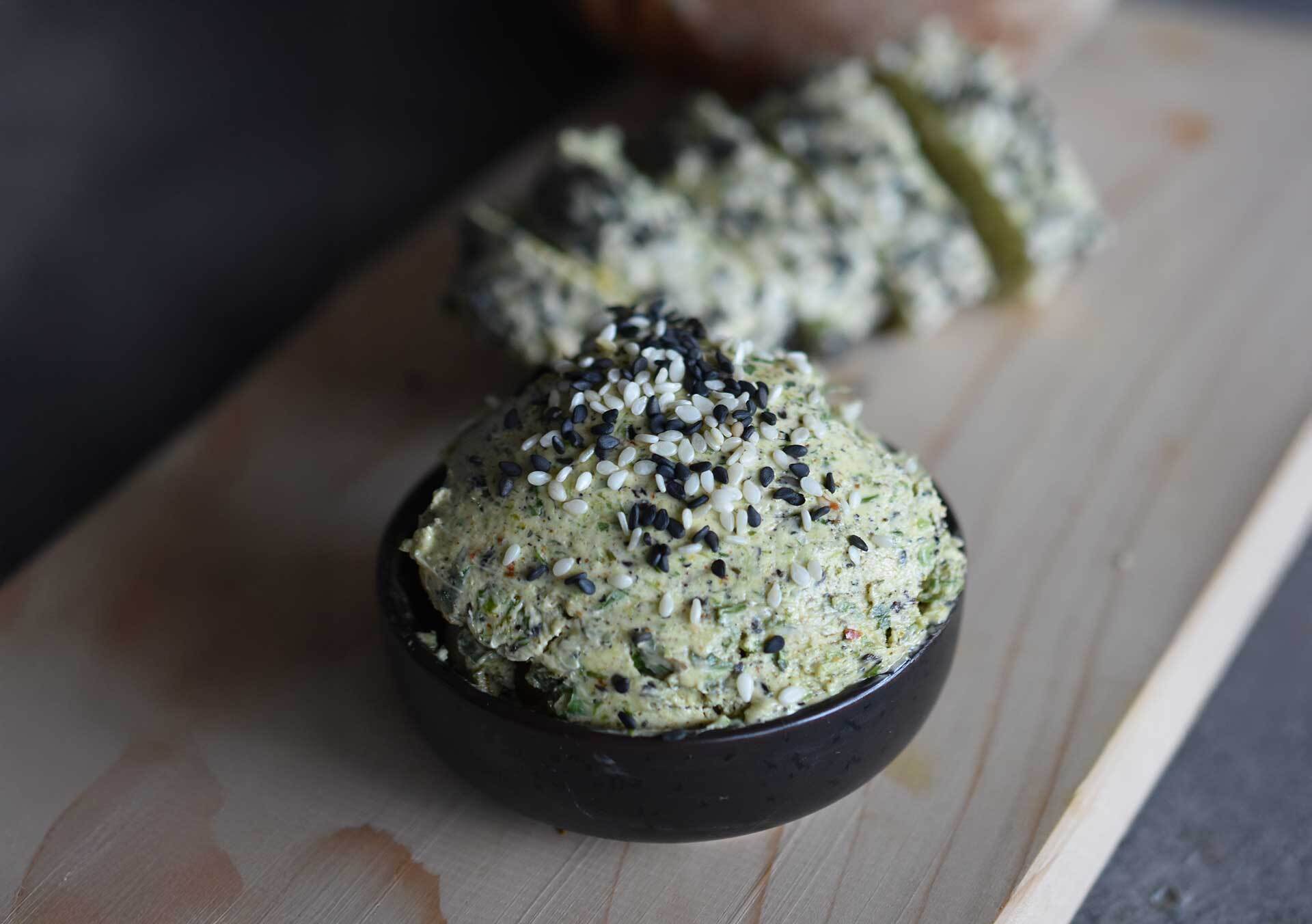 In einer kleinen schwarzen Schale liegt frisch angerührte Togarashi-Butter, bestreut mit schwarzem und weißem Sesam. Dahinter ist die angerollte Butterrolle auf einem Holzbrett zu sehen – Zubereitung im Stil der Gewürzmühle Rosenheim.