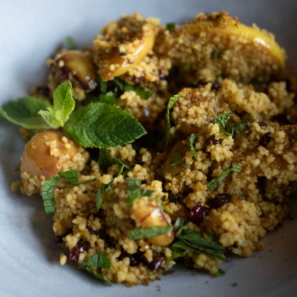 Fruchtiger Orangen-Couscous mit gebratenen Apfelspalten, Berberitzen und frischer Minze in einer hellen Keramikschüssel.