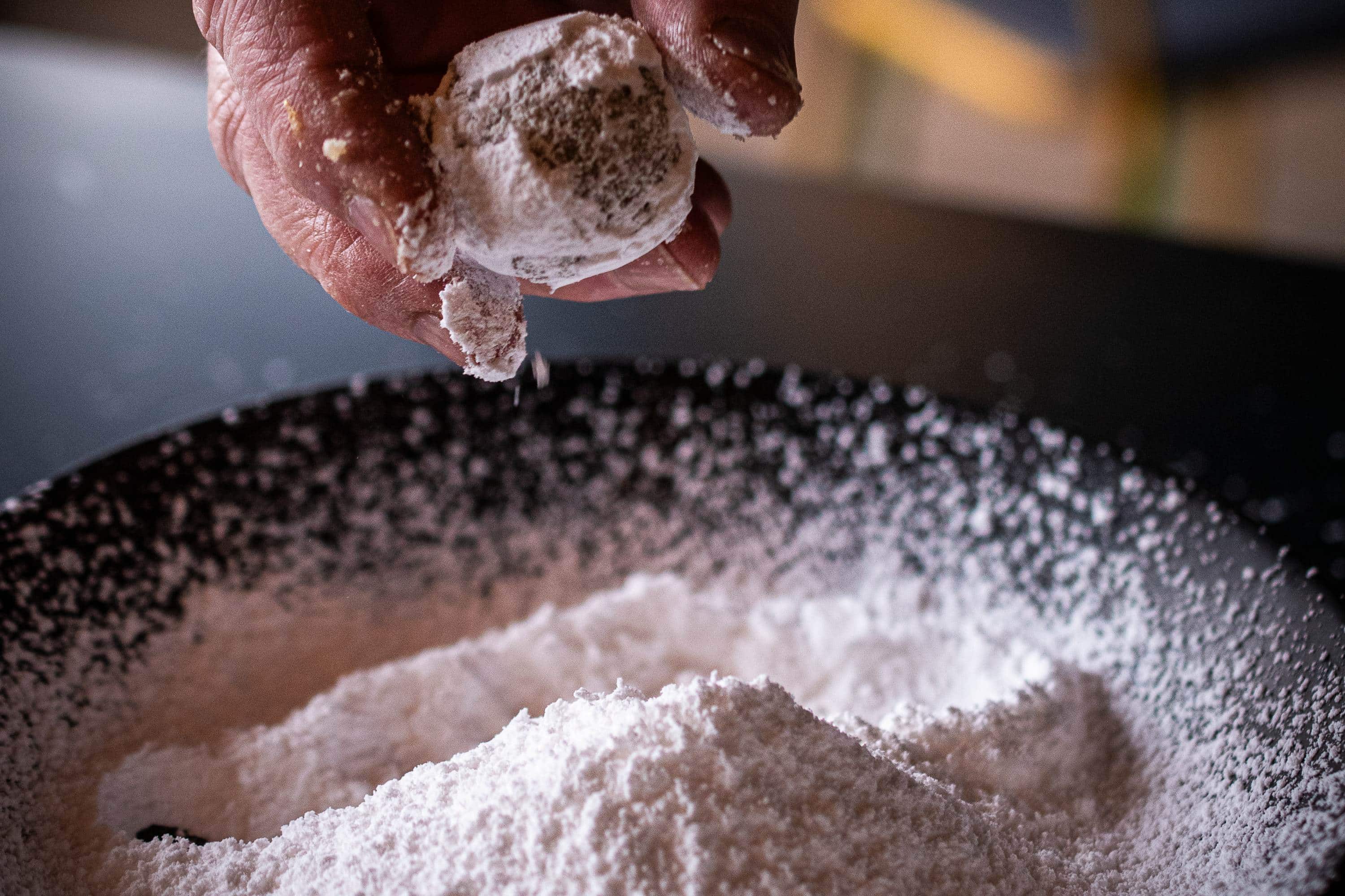 Hand wälzt ein Amaretti Morbidi in Puderzucker über einer dunklen Schale während der Zubereitung