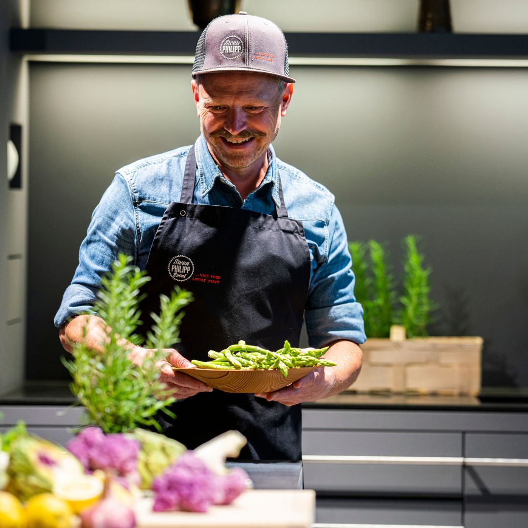 Koch in einer modernen Küche, lächelnd und mit einer Schale frischem grünem Spargel in den Händen. Im Vordergrund liegen buntes Gemüse und Kräuter bereit. Die Szene zeigt eine Zubereitungssituation, fotografiert für die Gewürzmühle Rosenheim.