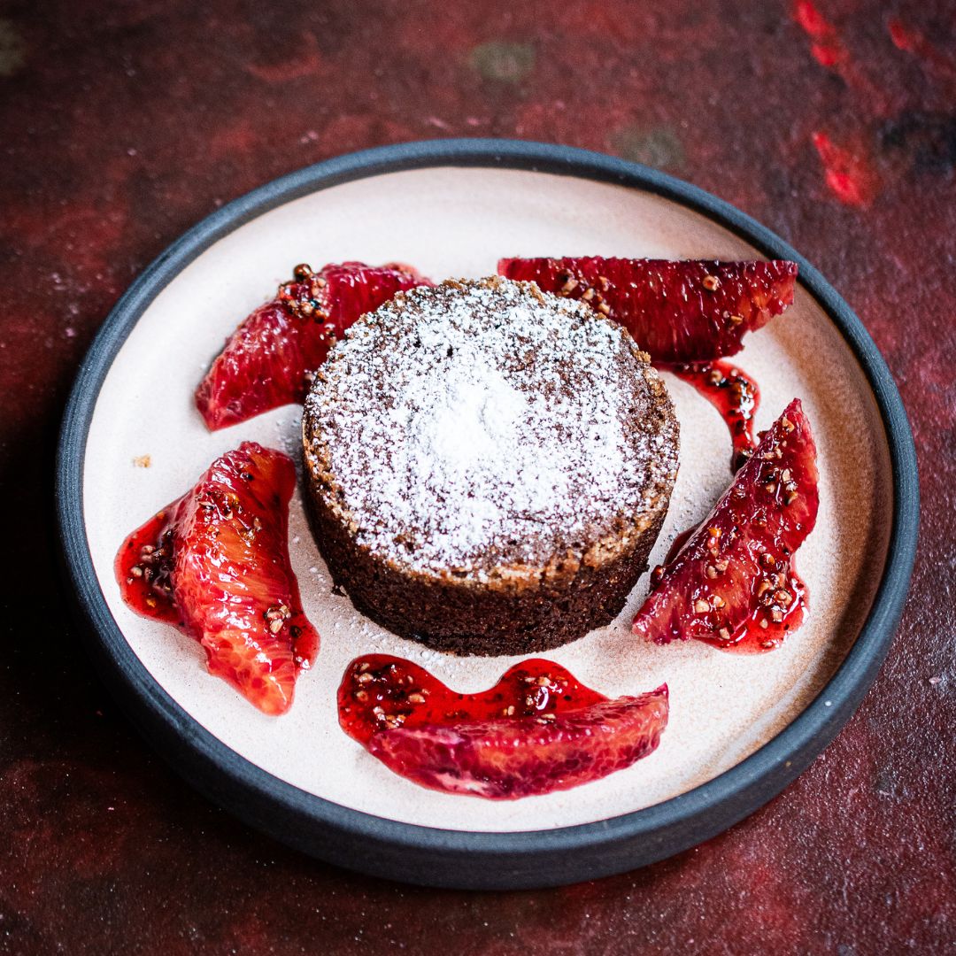 Ein rundes Schokoladenküchlein mit Puderzucker auf hellem Teller, umgeben von frischen Blutorangenfilets mit Gewürzkruste. Präsentiert im Kontext der Gewürzmühle Rosenheim.