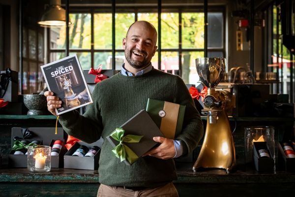 Simon Mendel der sein Kochbuch "SPICED" in der Hand hält, mit 2 weiteren Geschenken aus der Gewürzmühle Rosenheim; Im Hintergrund sind ebenfalls noch weitere verpackte Geschenke aus der Gewürzmühle Rosenheim