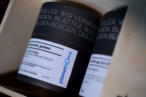 Nahaufnahme von zwei schwarzen Gewürzdosen der Gewürzmühle Rosenheim in einer hellen Geschenkbox. Die vordere Dose trägt ein weißes Etikett mit der blauen Aufschrift „planetHome“ und enthält geröstetes Bratkartoffelgewürz.