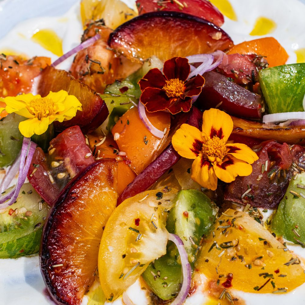 Ein bunter Tomaten-Pfirsich-Salat mit roten Zwiebeln, essbaren Blüten und italienischer Kräutermischung der Gewürzmühle Rosenheim, auf einer Feta-Creme.